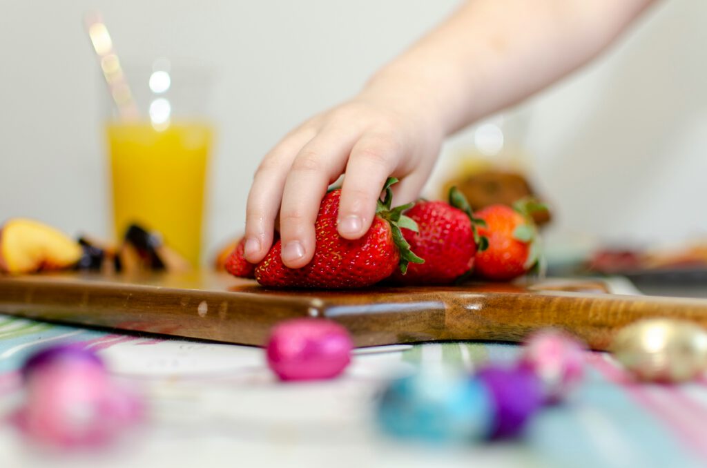 Why Texture Matters: The Secret Power of Pectin in Healthy Gummies 1 A child’s hand reaching for fresh strawberries on a wooden board, with a glass of orange juice and colorful treats in the background, highlighting the importance of taste and texture in kids’ snacks.